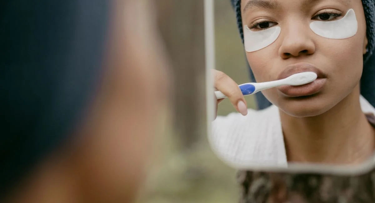 A black woman in a bathroom brushes her teeth while wearing cosmetic eye patches in front of a mirror.