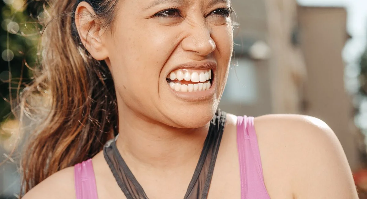 A woman in a pink top smiles brightly outdoors, showcasing an expressive face.