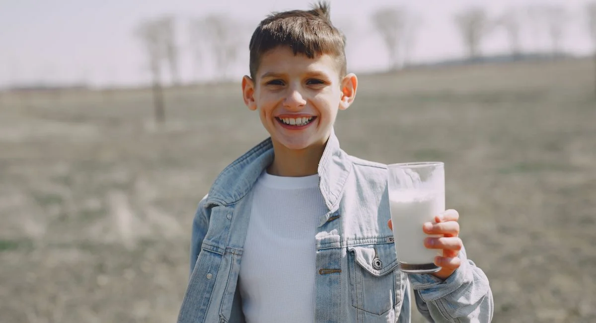 Smiling little child in casual outfit with glass of milk in hand smiling and looking at camera in countryside in field
