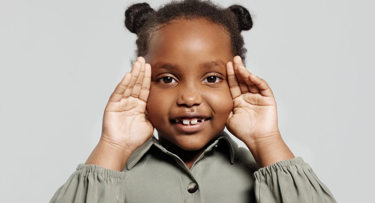 Adorable child with curly hair smiling in a studio portrait, hands framing face, exuding joy and warmth.