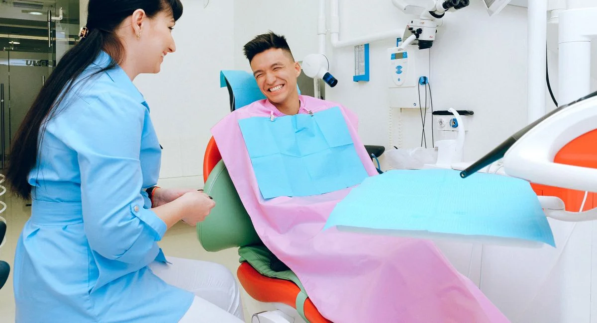 Happy ethnic guy sitting in dental chair and smiling while talking with female dentist in uniform before treatment in modern 