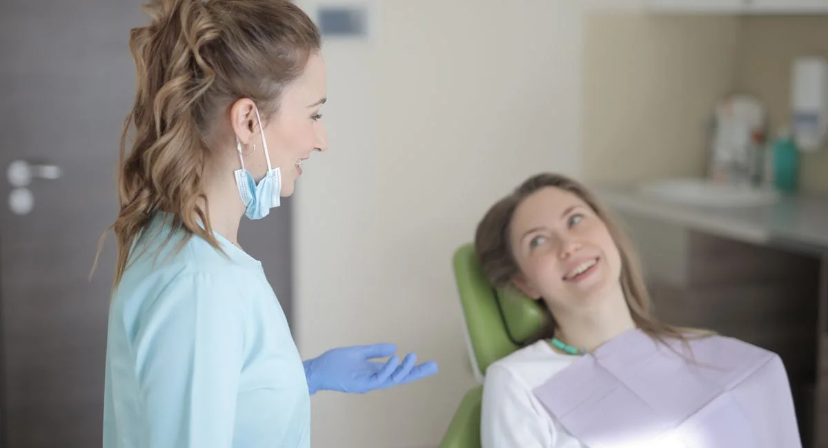 Positive young female dentist in latex gloves and uniform talking with young patient while sitting near chair and preparing f