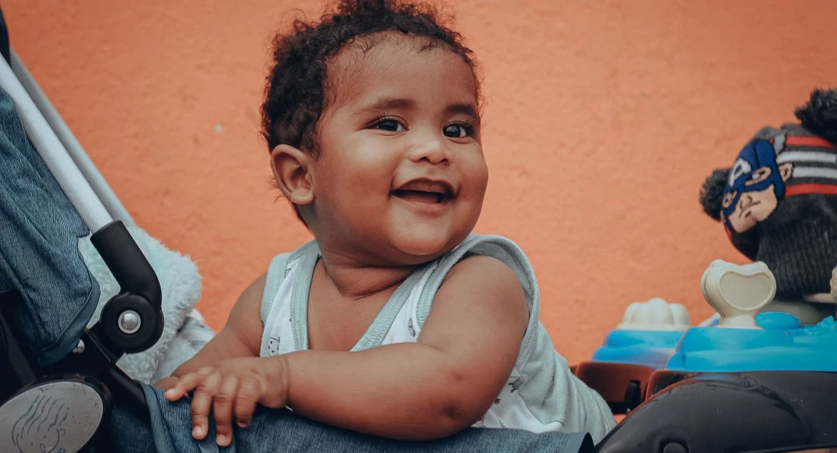 Adorable baby sitting and smiling in a stroller with a vibrant orange background.