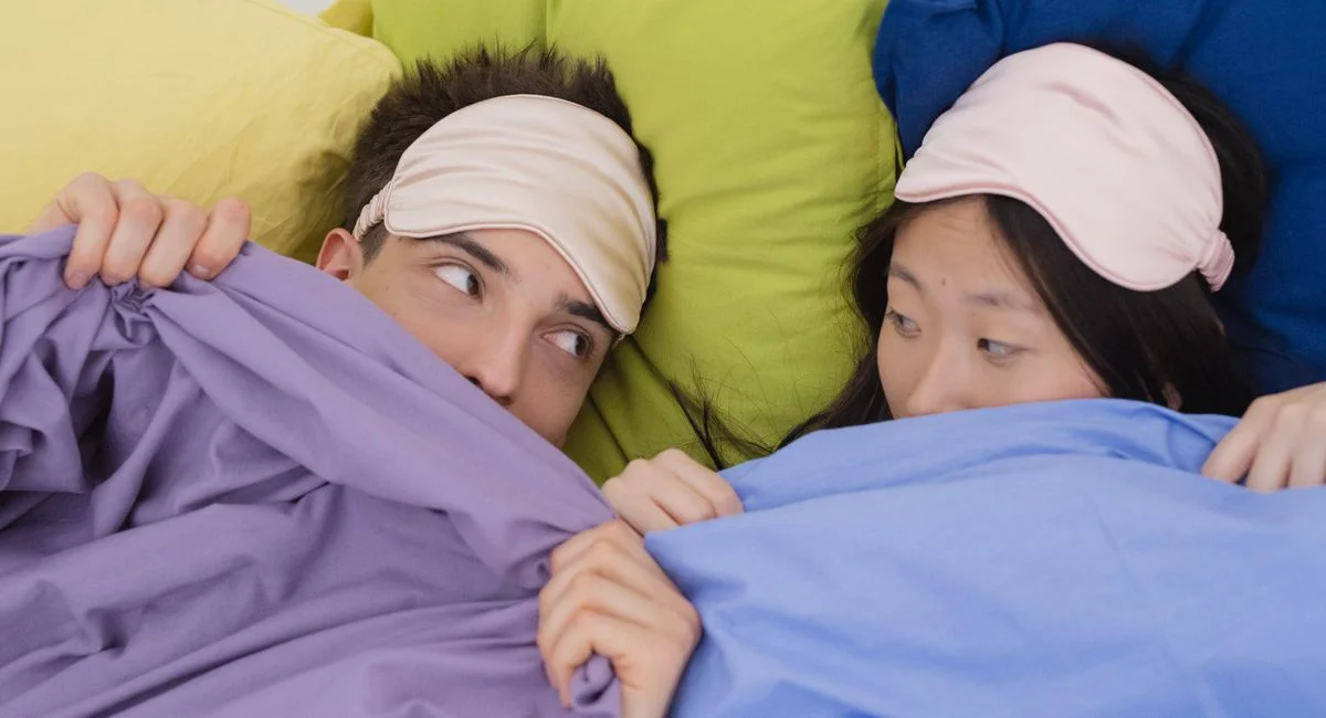 An interracial couple wearing sleep masks lying down on colorful pillows, portraying relaxation and rest.