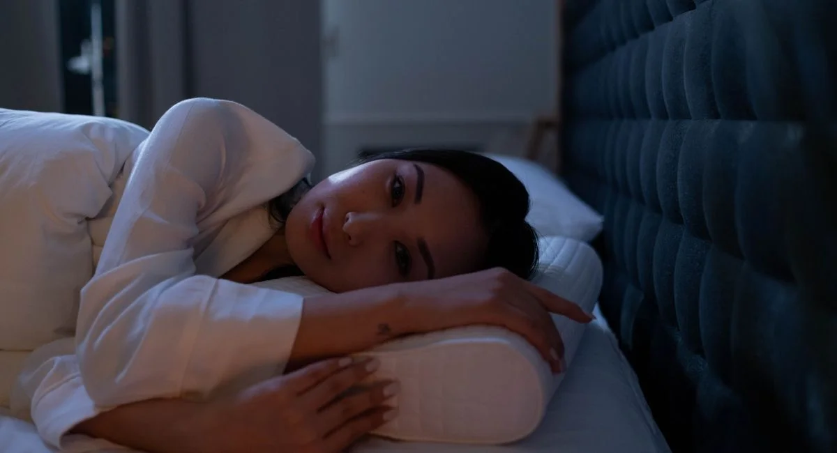 Dark bedroom scene of an Asian woman with insomnia lying on her side, looking thoughtful.