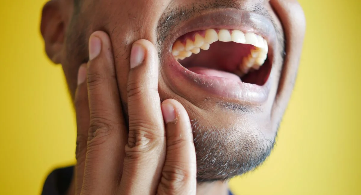Close-up of a man holding his cheek, displaying a wide open mouth against a yellow background.