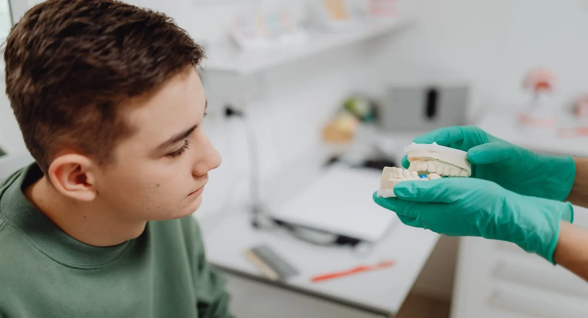 Teenager attending a dental consultation, examining a dental model held by a dentist wearing green gloves.
