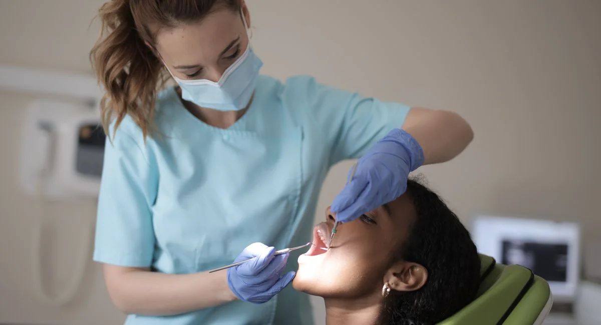 Dentist in blue scrubs and mask examines patient's teeth in clinic setting.