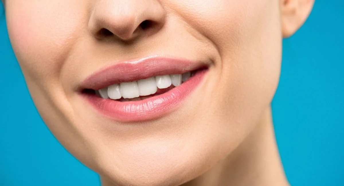 A close-up image of a woman's smile with pink lipstick and white teeth against a blue background.